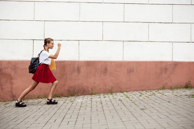 Young beautiful female student in glasses walking down street, holding folders