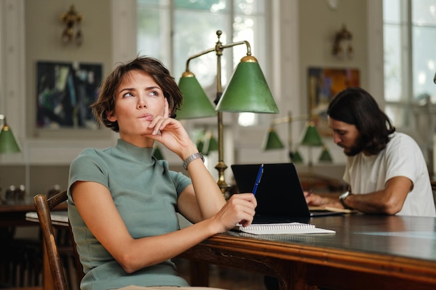 Young beautiful female student with notepad dreaming during study in library of university