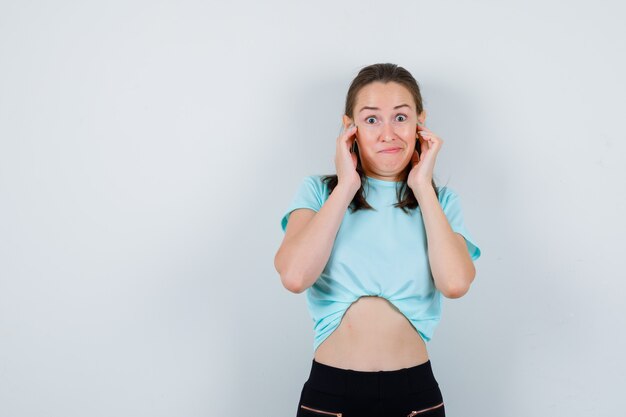 Young beautiful female in t-shirt plugging ears with fingers and looking wondered , front view.