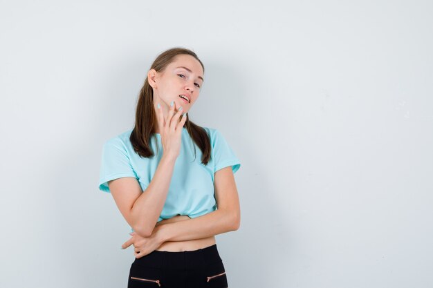 あごに指を置いてポーズをとって魅力的に見えるTシャツの若い美しい女性、正面図。