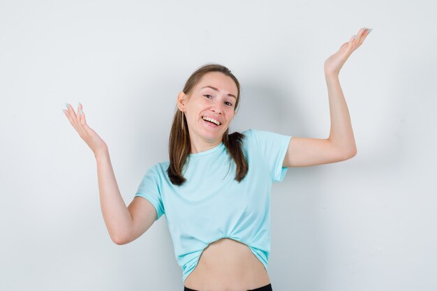 Young beautiful female in t-shirt raising hands in questioning manner and looking blissful , front view.