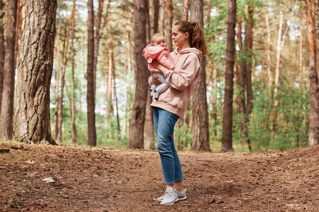 Young beautiful female with brown hair and ponytail holding small girl in hands, walking in park or forest together