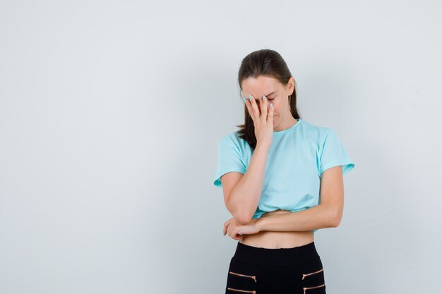 Young beautiful female with hand on face in t-shirt, pants and looking disappointed , front view.