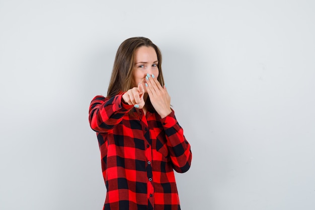 Young beautiful female with hand on mouth, pointing at camera in casual shirt and looking glad. front view.