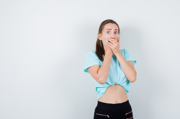 Young beautiful female with hands on mouth in t-shirt, pants and looking scared. front view.