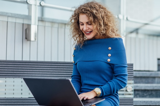 Free photo young beautiful female with a laptop sits on a bench in the business part of the city. young beautiful woman, freelancer, works on the laptop in summer wear blue dress.