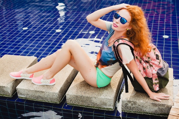 Young beautiful ginger woman sitting at pool with backpack, relaxed, happy, summer, cool hipster outfit, shorts, t-shirt, sneakers, sunglasses