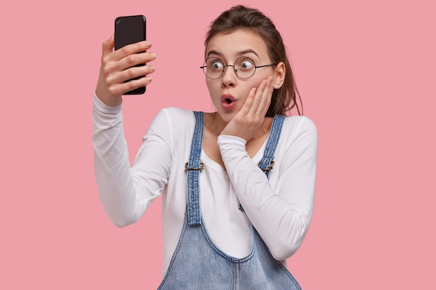 Young beautiful girl amazed by recieved email, makes video call for sharing news with friend on distance, connected to high speed internet holds cellular