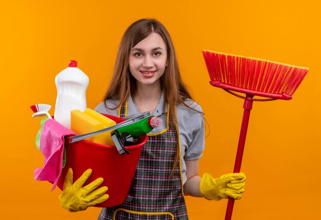 Young beautiful girl in apron holding bucket with cleaning tools and mop smiling cheerfully 