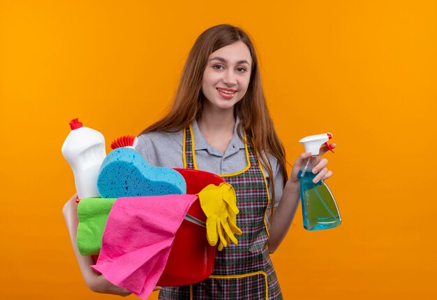 Young beautiful girl in apron holding bucket with cleaning tools and spray smiling cheerfully 