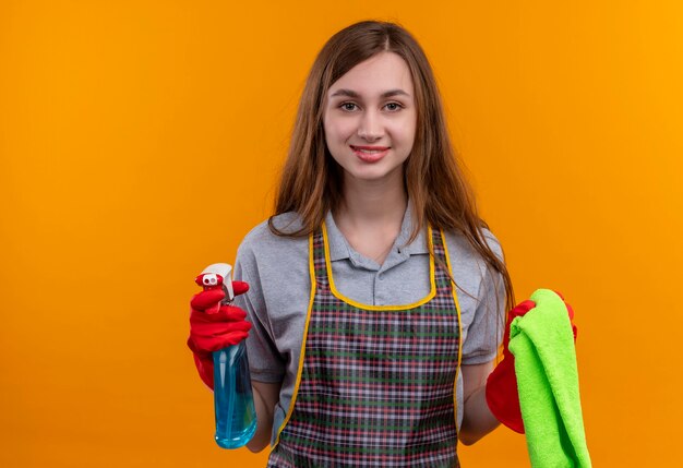 Young beautiful girl in apron holding cleaning spray and rug looking at camera smiling , ready for cleaning 