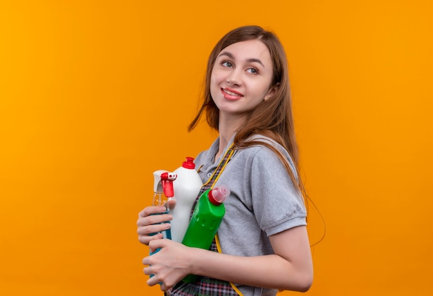 Young beautiful girl in apron holding cleaning supplies looking aside smiling friendly, ready for cleaning 