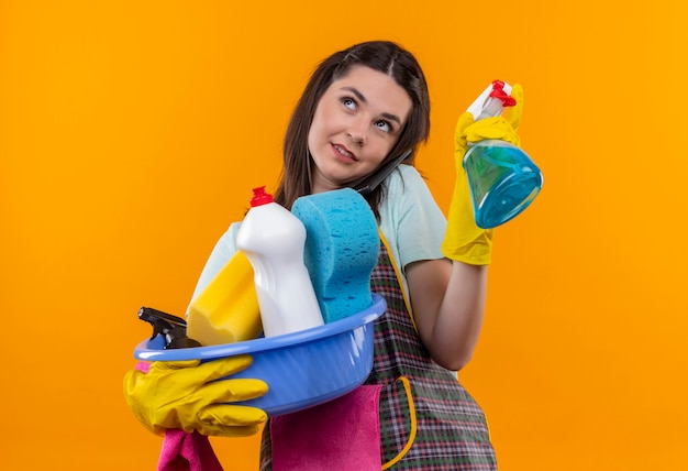 Free photo young beautiful girl in apron and rubber gloves holding basin with cleaning tools and cleaning spray looking up smiling with happy face