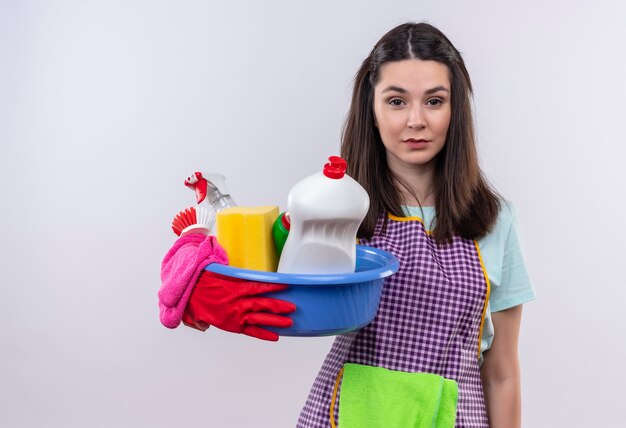 Young beautiful girl in apron and rubber gloves holding basin with cleaning tools looking at camera tired and bored 