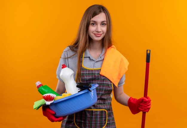 Young beautiful girl in apron and rubber gloves holding basin with cleaning tools and mop smiling confident 