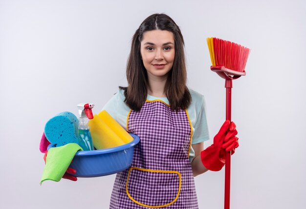 Young beautiful girl in apron and rubber gloves holding basin with cleaning tools and mop smiling confident 
