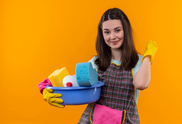 Young beautiful girl in apron and rubber gloves holding basin with cleaning tools smiling friendly looking at camera pointing back
