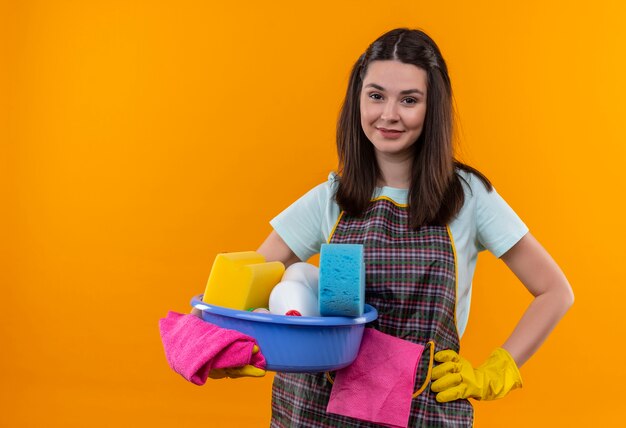 Young beautiful girl in apron and rubber gloves holding basin with cleaning tools smiling friendly looking at camera