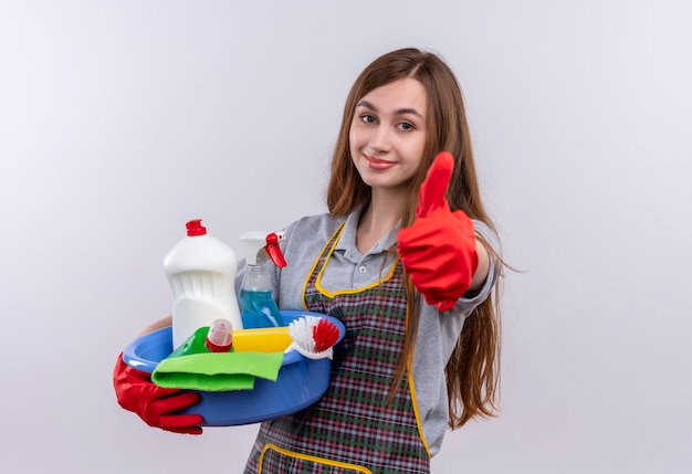 Young beautiful girl in apron and rubber gloves holding basin with cleaning tools smiling showing thumbs up 