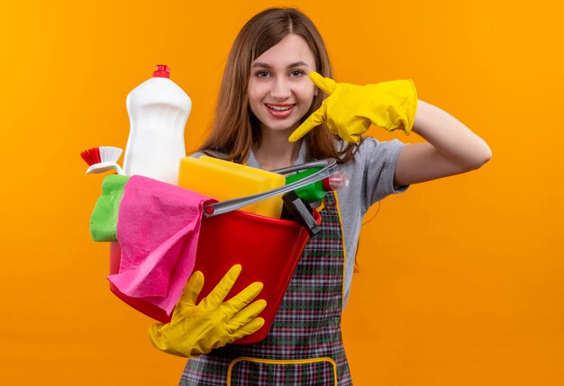 Young beautiful girl in apron and rubber gloves holding bucket with cleaning tools pointing with finger to it smiling cheerfully 