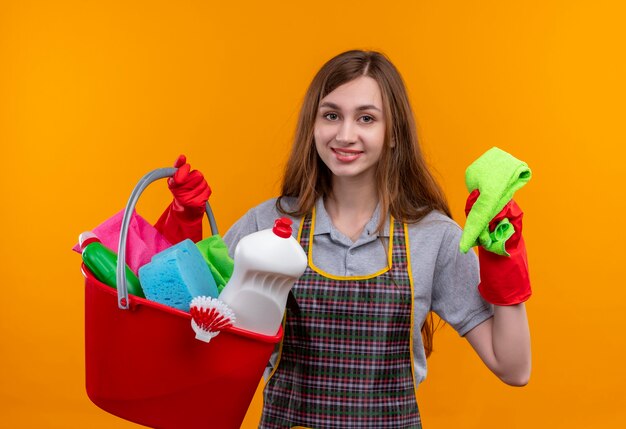 Giovane bella ragazza in grembiule e guanti di gomma che tiene secchio con strumenti di pulizia e tappeto sorridendo allegramente guardando la telecamera, pronta per la pulizia