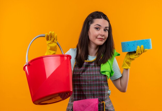 Young beautiful girl in apron and rubber gloves holding buclet with sponge smiling confident