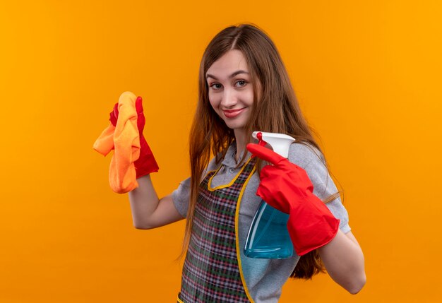 エプロンとゴム手袋で若い美しい少女が掃除スプレーと敷物を持ってカメラを見てポジティブで幸せな笑顔、掃除の準備ができて