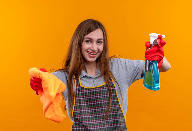 エプロンとゴム手袋で若い美しい少女が掃除スプレーと敷物を持ってカメラの笑顔を見て、掃除の準備ができて