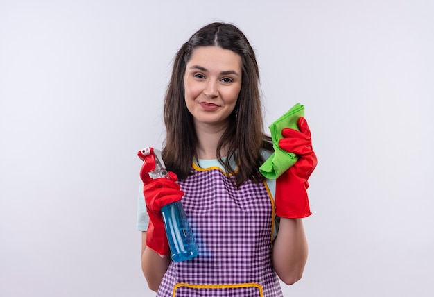 Foto gratuita giovane bella ragazza in grembiule e guanti di gomma che tengono spray per la pulizia e tappeto sorridente fiducioso, pronto per la pulizia