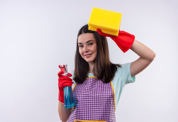 Young beautiful girl in apron and rubber gloves holding cleaning spray and sponge looking at camera smiling confident, ready for cleaning 