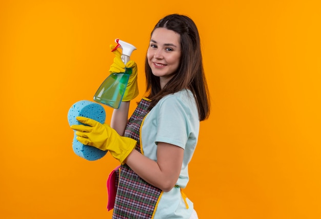 Free photo young beautiful girl in apron and rubber gloves holding cleaning spray and sponge looking at camera smiling with happy face standing sideways over orange background