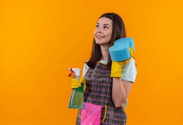 Young beautiful girl in apron and rubber gloves holding cleaning spray and sponge looking up smiling confident