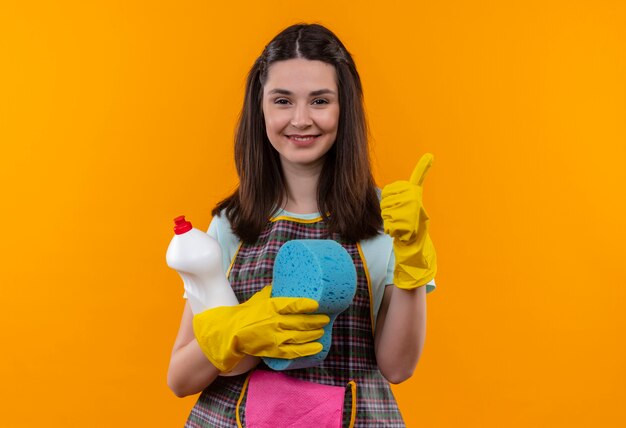 Young beautiful girl in apron and rubber gloves holding cleaning suplies and sponge looking at camera smiling cheerfully showing thumbs up