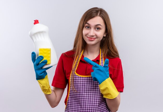 Молодая красивая девушка в фартуке и резиновых перчатках держит чистящие средства, улыбаясь, указывая на нее указательным пальцем