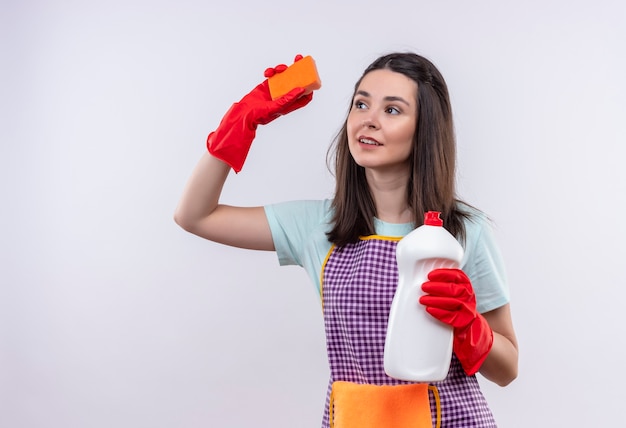 Young beautiful girl in apron and rubber gloves holding cleaning supplies and sponge smiling confident, going to clean 