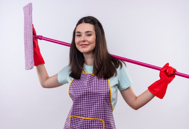 Young beautiful girl in apron and rubber gloves holding mop smiling confident looking at camera 