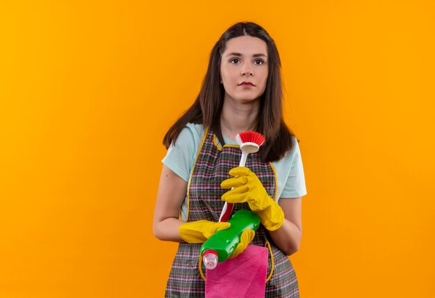 Young beautiful girl in apron and rubber gloves holding scrubbing brush and cleaning supp looking aside with serious facelies