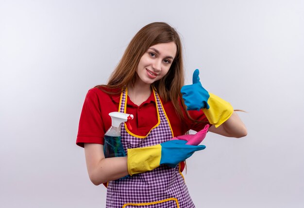 Young beautiful girl in apron and rubber gloves holdingcleaning supplies smiling cheerfully showing thumbs up 