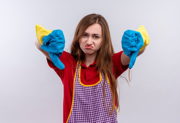 Giovane bella ragazza in grembiule e guanti di gomma che guarda l'obbiettivo scontento che mostra i pollici verso il basso con entrambe le mani