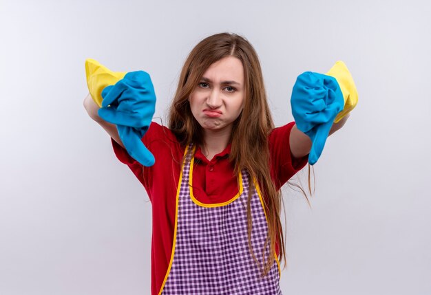 Young beautiful girl in apron and rubber gloves  looking at camera displeased showing thumbs down with both hands 