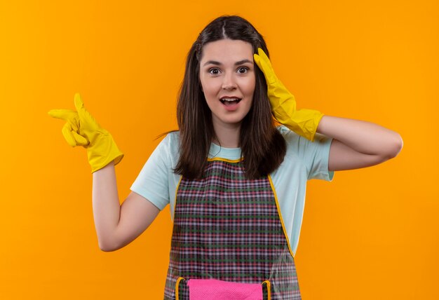 Young beautiful girl in apron and rubber gloves looking surprised and amazed pointing with finger to the side