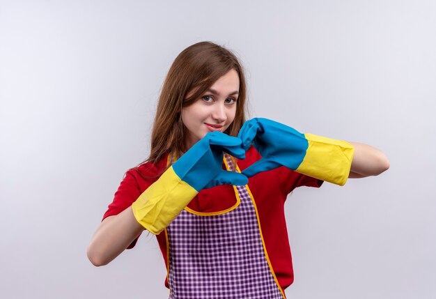 Young beautiful girl in apron and rubber gloves  making heart gesture with fingers over chest smiling 