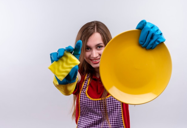 Young beautiful girl in apron and rubber gloves showing plate and  sponge, smiling cheerfully 