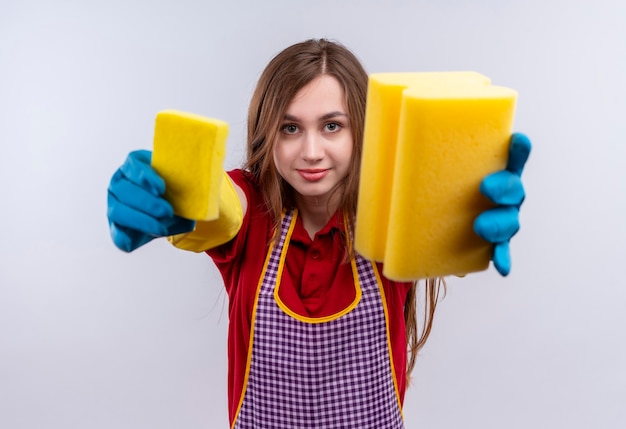 Young beautiful girl in apron and rubber gloves  showing sponges to camera smiling 