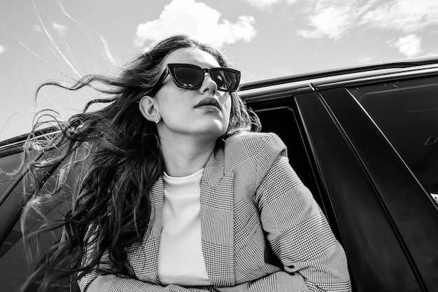 A young, beautiful girl in a black car looks out of the window. a stylish girl with glasses rides in a car leaning out of the window. black and white photo Premium Photo