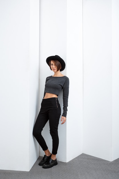 Foto gratuita giovane bella ragazza in cappello nero che posa sopra la parete bianca