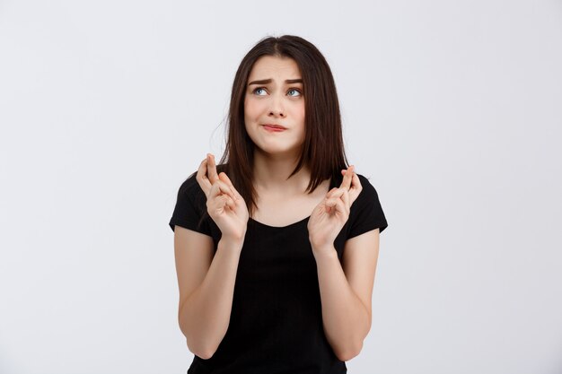 Young beautiful girl in black t-shirt posing with hopefully crossed fingers  over white wall