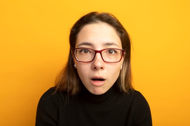 Young beautiful girl in a black turtleneck and glasses amazed and surprised 