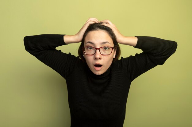 Young beautiful girl in a black turtleneck and glasses bainf confused and surprised with hands on her head 