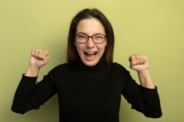 Young beautiful girl in a black turtleneck and glasses clenching fists happy and excited 
